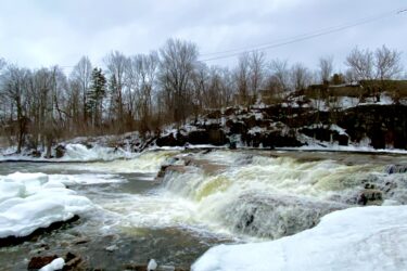A winter landscape featuring a cascading waterfall surrounded by ice and snow. The scene includes rocky outcrops and bare trees in the background under a cloudy sky. Almonte Riverside Trail mountain bike trail.