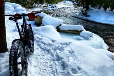 A fat bike resting on a snowy trail alongside a flowing river, surrounded by evergreen trees. The path is covered in fresh snow, with visible tire tracks leading into the winter landscape. Inglis falls West rock mountain bike trail.