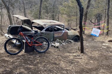 An old, rusted car with its hood open is surrounded by trees in a forested area. In the foreground, a mountain bike is leaning against a wooden crate. A sign nearby warns that the trail is under construction and indicates no entry, with caution tape marking the area. Cedar Niles Park mountain bike trail.
