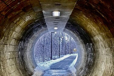 A dimly lit concrete tunnel leading to a snowy pathway, with lights along the ceiling and trees visible in the background. The path is partially covered in snow, and a sign can be seen in the distance. Springbank park mountain bike trail.