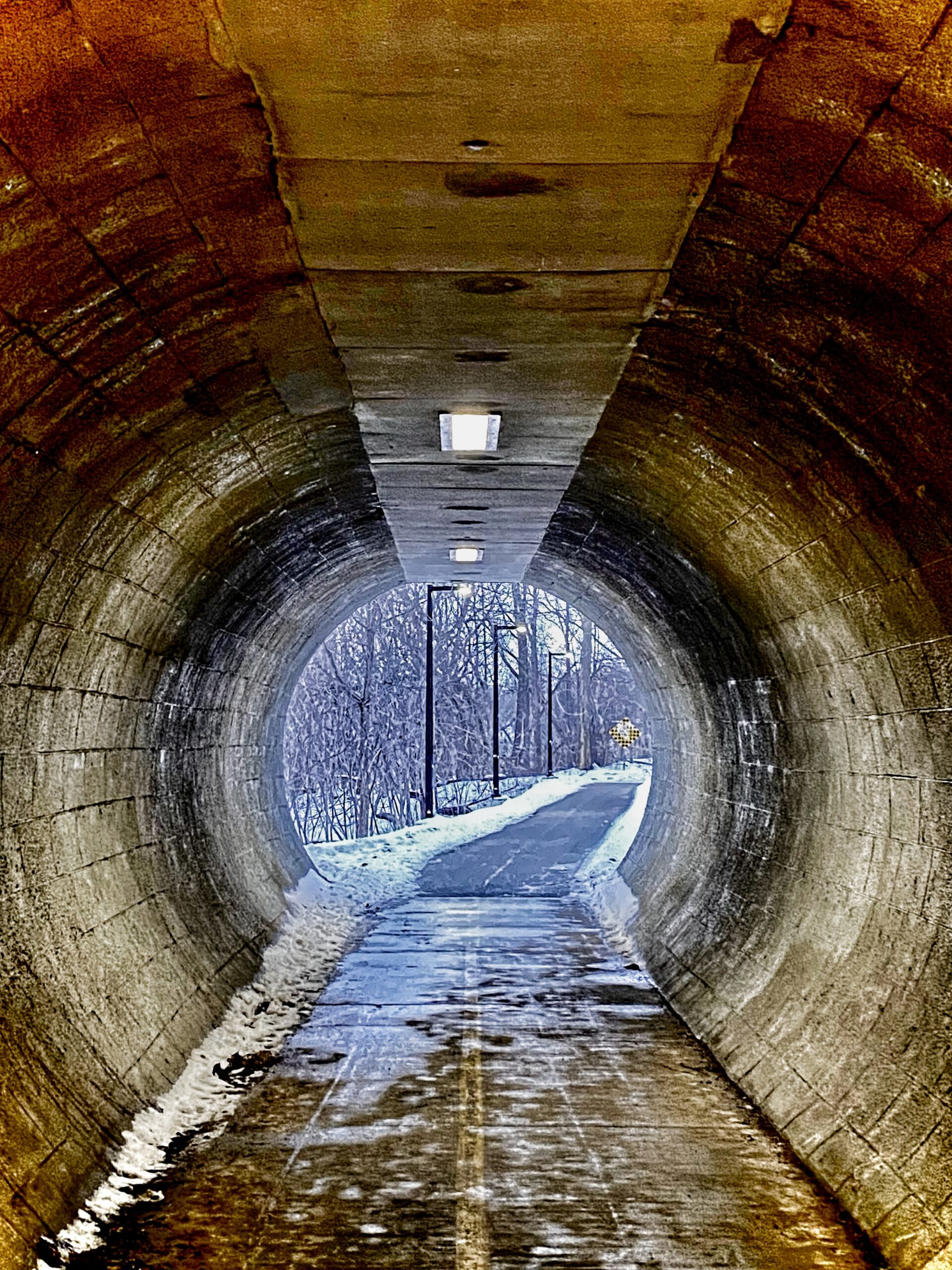 A dimly lit concrete tunnel leading to a snowy pathway, with lights along the ceiling and trees visible in the background. The path is partially covered in snow, and a sign can be seen in the distance. Springbank park mountain bike trail.