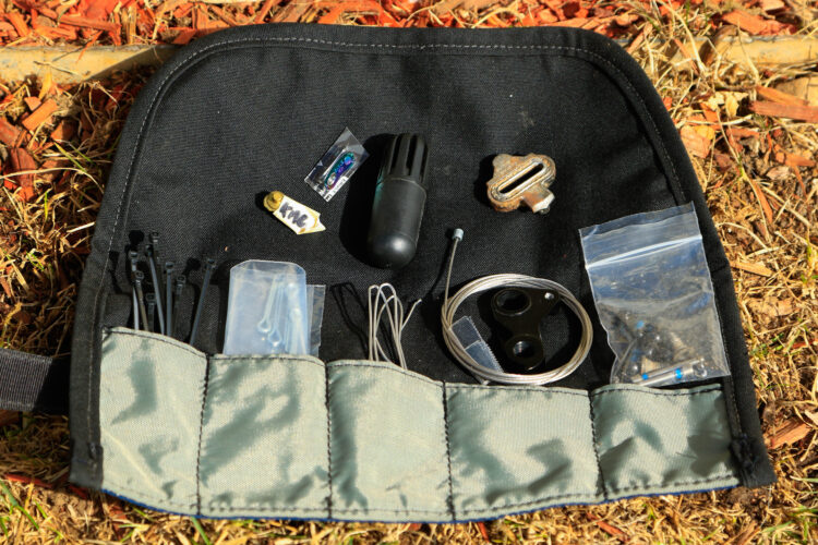 A close-up view of a black tool pouch opened to display various small tools and items inside, including zip ties, a black cylindrical object, small plastic bags containing components, metal pieces, and wires, all neatly organized. The pouch is placed on grassy ground with wood chips visible in the background.