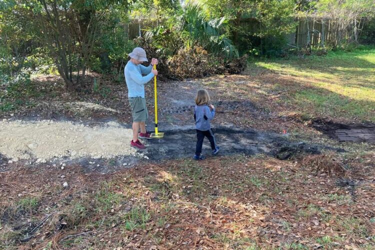 A person using a yellow digging tool in a yard while a child observes nearby. The area shows a path with light-colored gravel, surrounded by grass and trees.