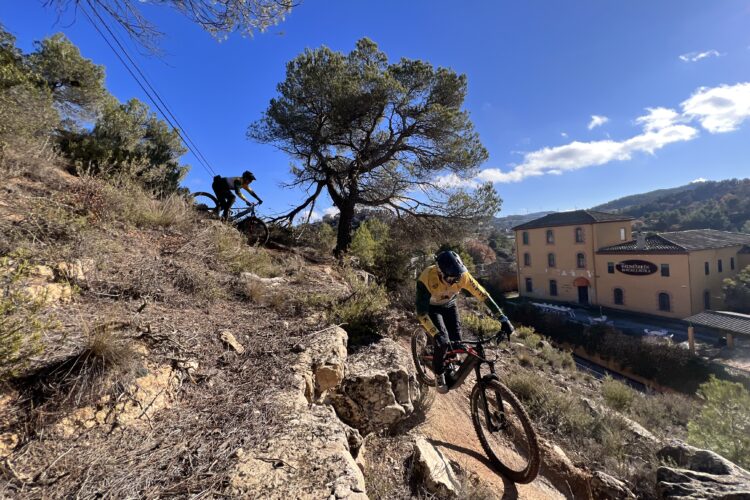 A mountain biker navigating a rocky trail surrounded by trees, with a building in the background under a clear blue sky. The rider is wearing a helmet and a bright yellow and green jersey, emphasizing a dynamic outdoor sports scene.