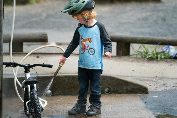 A young child wearing a bicycle helmet and a colorful dinosaur-themed shirt is spraying water on a bike with a hose at an outdoor area. The child is focused on cleaning the bike, with gravel and greenery visible in the background. The scene captures a moment of playful activity and outdoor fun.