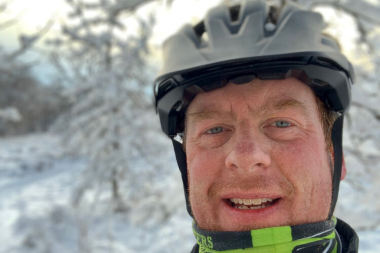 A person wearing a black helmet and a green and black neck warmer smiles for a selfie in a snowy landscape with trees in the background. The scene captures a bright winter day.