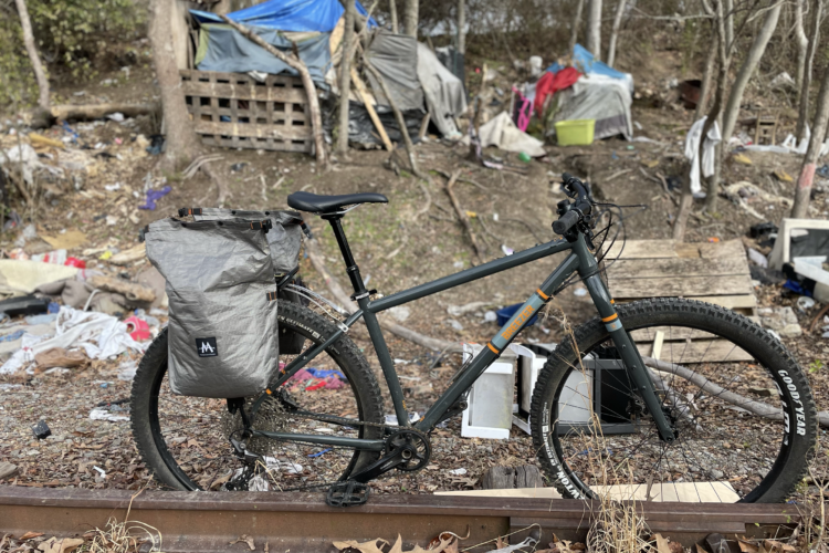 A bicycle with a grey bag attached to it is positioned on a railway track in the foreground. Behind the bike, there is a makeshift campsite with large tarps, debris, and scattered items among trees, highlighting a neglected area.