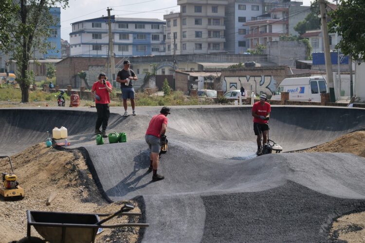 Four workers are constructing a skate park, focused on shaping a concrete surface. Two individuals are in red shirts, while one is using a trowel on the ground. Green containers and equipment are scattered around, and buildings can be seen in the background under a partly cloudy sky.