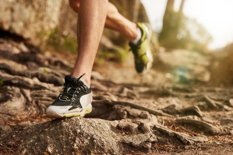 Close-up of a runner's foot in a black and white athletic shoe stepping over rocky terrain, with sunlight filtering through trees in the background. The image captures the dynamic movement of trail running in a natural setting.