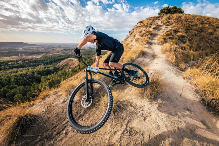 A mountain biker in a helmet skillfully maneuvers down a rugged hill path surrounded by dry grass and trees, with a scenic view of the valley and cloudy sky in the background.