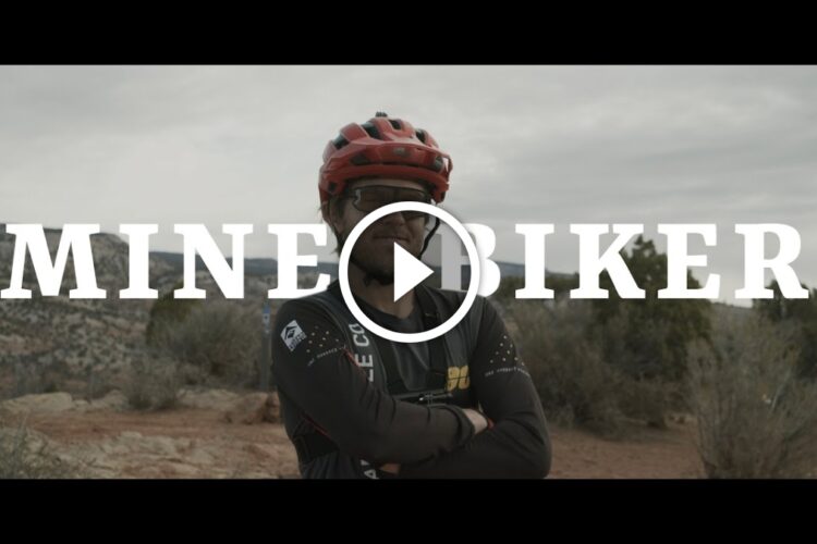A person wearing a red helmet and a cycling jersey stands confidently with their arms crossed in a rugged outdoor mountain setting. The text "MINE BIKER" is prominently displayed across the image, with a play button symbol in the center.