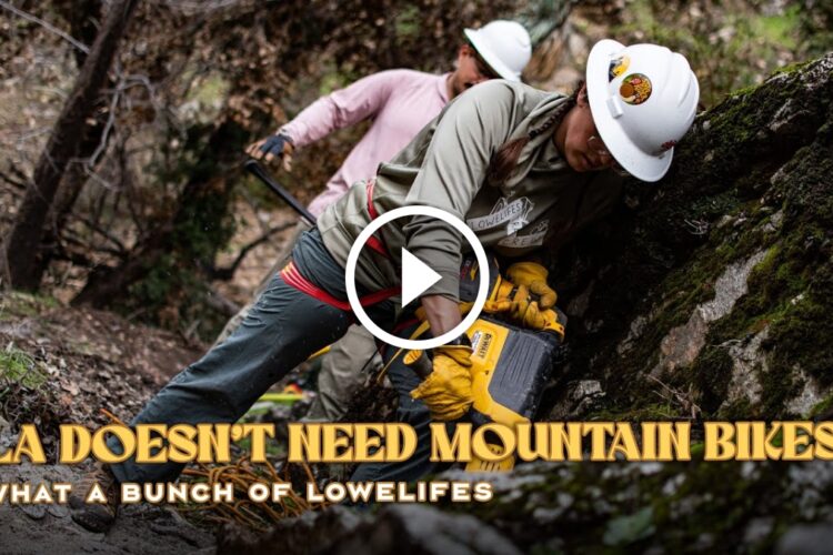 A group of individuals wearing hard hats and work attire are engaged in trail maintenance in a forested area. One person is using a power tool to work on the ground while others assist nearby. The scene captures the effort of the team in maintaining the trails, promoting outdoor recreation. The image includes overlay text that reads "LA Doesn't Need Mountain Bikes: What a Bunch of Lowlifes."