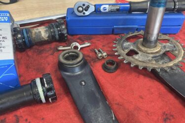Close-up image of bicycle components on a red workbench. The scene includes a bottom bracket, a chainring with visible dirt, several tools including a ratchet and a torque wrench, and a few loose screws. The background features a blue tool case and an assortment of bicycle parts, highlighting a bicycle maintenance or repair activity.