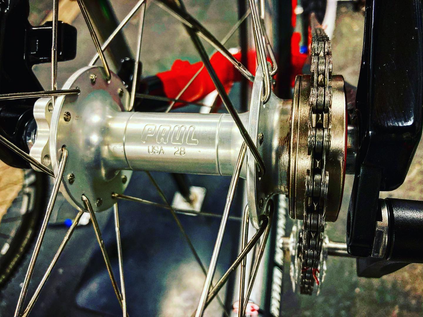 Close-up of a bicycle rear hub featuring the brand name "Paul" and the designation "USA 28," with visible spokes, a portion of the chain, and part of the bike frame in the background.