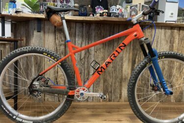 A vibrant orange Marin mountain bike with thick tires and a blue front fork is displayed in a rustic shop environment. The bike features a single gear setup, a sleek frame design, and a brown saddle bag attached to the seat. The wooden walls of the shop and various accessories in the background provide a cozy atmosphere.