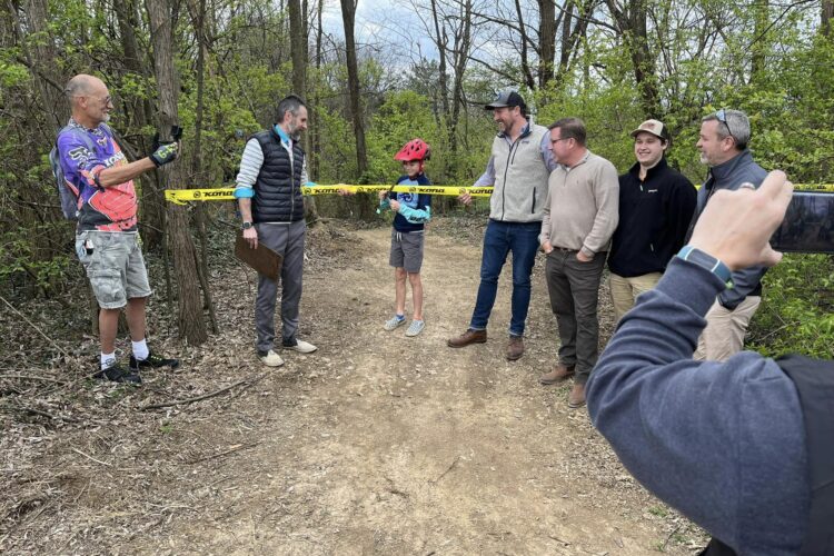 A group of six people stands on a dirt path in a wooded area, gathered around a yellow ribbon barrier. One person, an older man in a colorful shirt, is taking a photo with a smartphone. A young boy in a helmet and safety gear is positioned in front of the ribbon, while others look on, smiling. The scene suggests a trail opening or cycling event.
