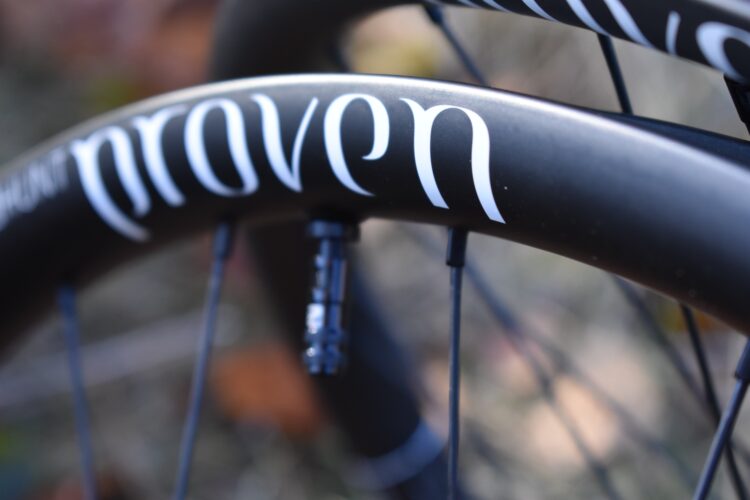 Close-up of a bicycle wheel rim featuring the word "proven" in bold white lettering against a black background. The image captures details like the valve and spokes, with a blurred natural environment in the background.