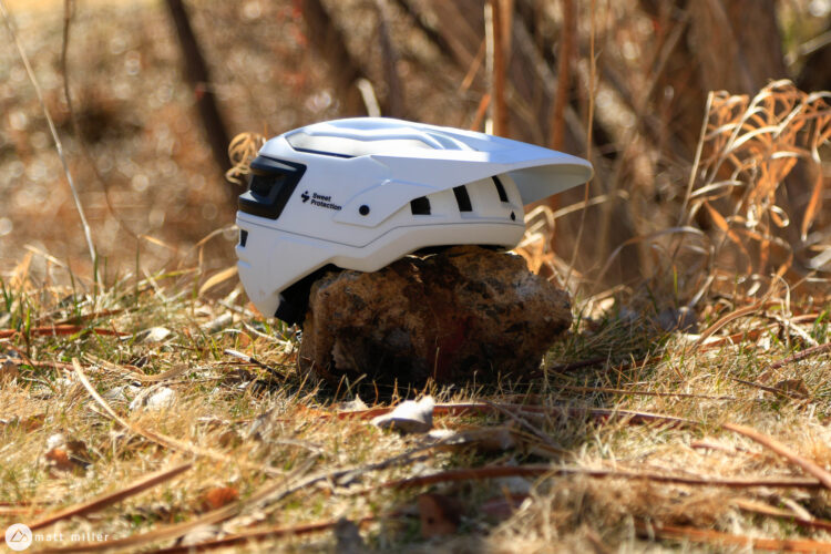 A white bicycle helmet resting on a rock, surrounded by grass and dry foliage. Soft sunlight filters through the trees in the background, creating a natural outdoor setting.