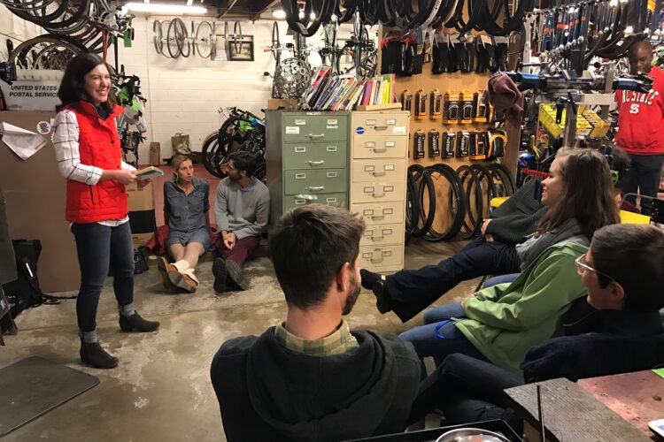 A group of people is seated in a bike shop during a presentation. A woman in a red vest stands in the foreground, smiling and holding a notebook while speaking to the audience. The background features bicycles and bike parts on the walls, along with shelves and storage units filled with tools and accessories. The audience consists of a diverse group of individuals, some sitting cross-legged on the floor, engaged and listening attentively.