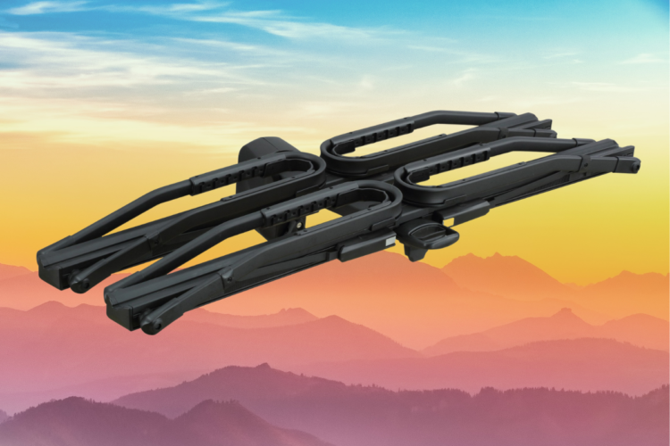 A black, multi-functional folding tool or device with curved arms, set against a gradient background of orange and blue skies with mountain silhouettes.
