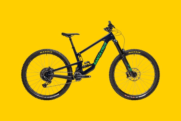 A black and teal mountain bike positioned against a bright yellow background, showcasing its sturdy frame and large knobby tires designed for off-road performance. The bike features a dual suspension system and a prominent brand logo on the frame.