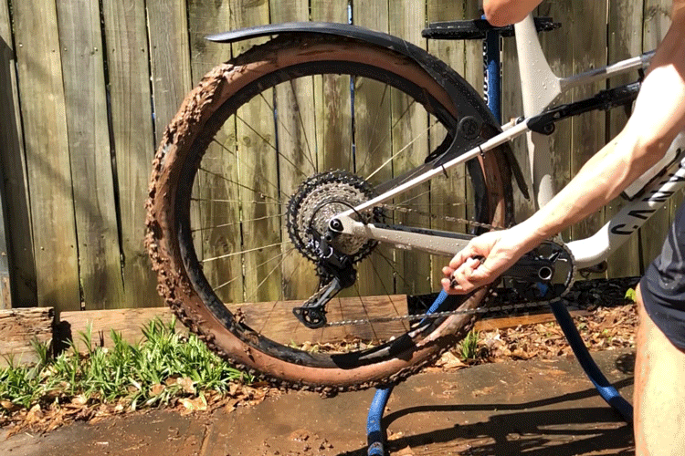 A person cleaning the rear wheel and drivetrain of a mountain bike in a sunny outdoor setting, with mud visibly caked on the tire and components. The bike is propped up on a stand, and the background features a wooden fence and grassy area.