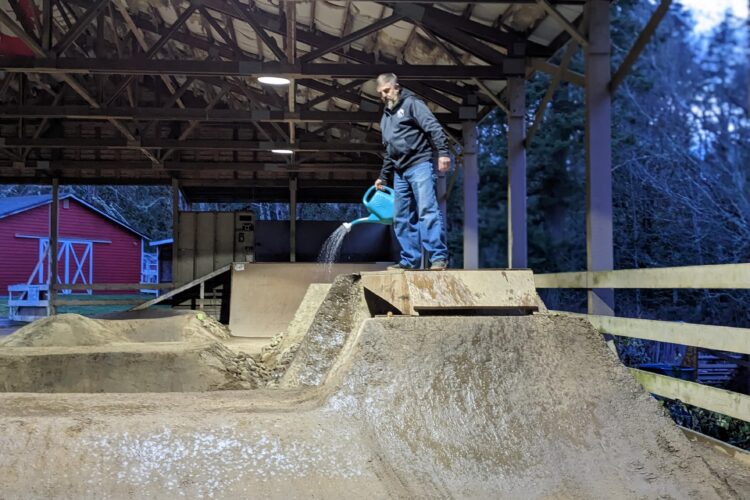 A person standing on a dirt ramp inside a covered area, pouring water from a blue watering can onto the dirt surface. The background features additional dirt jumps and a red barn outside. The scene is illuminated by overhead lights, indicating early evening.