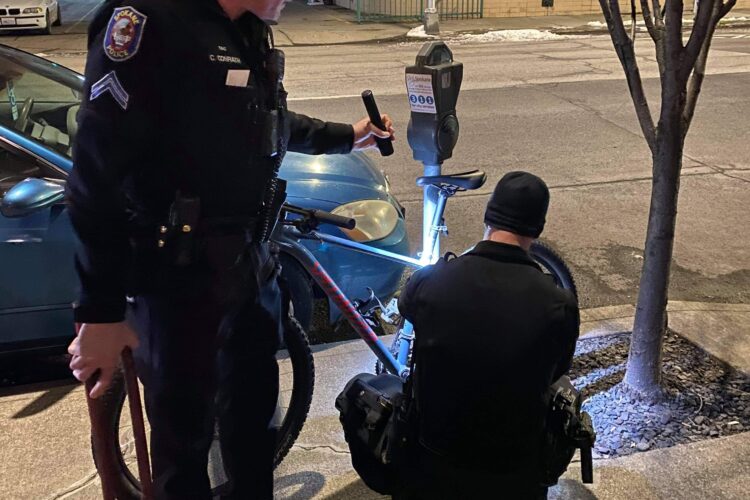 Two police officers are standing next to a parked car at night, with one officer using a flashlight to inspect a parking meter while the other officer crouches nearby. The scene is illuminated by city lights, and there are trees and a building in the background.