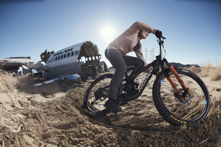 A cyclist riding a mountain bike through sandy terrain near an abandoned airplane, with the sun shining in the background. The scene conveys a sense of adventure and exploration in a rugged environment.