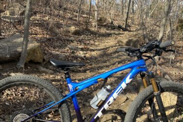 A blue mountain bike resting on a rocky trail surrounded by bare trees and a clear blue sky. The terrain is uneven with scattered leaves and rocks, indicating an outdoor biking path in a natural setting. Perry Lake Bike Trail mountain bike trail.
