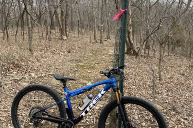 A blue and black mountain bike rests on a rocky path in a wooded area during late autumn or early winter. In the background, a signpost labeled "Great White" is attached to a tree, adorned with a pink ribbon. The ground is covered with dry leaves, and the trees are bare, indicating the season. Perry Lake Bike Trail mountain bike trail.