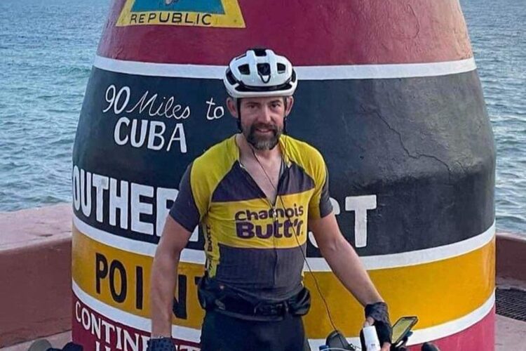A cyclist wearing a yellow and gray jersey stands next to a large colorful buoy at the Southernmost Point in Key West, Florida. The buoy features a conch shell design and text indicating the distance to Cuba. The background includes the ocean and a partly cloudy sky. The cyclist's bike is equipped with several bags for gear and supplies.