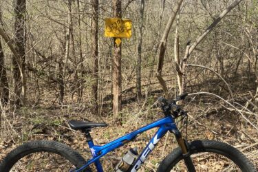 A blue mountain bike resting on the ground near a trail in a wooded area. A yellow caution sign indicating "CHURCH CAB" is visible in the background, surrounded by bare trees and scattered leaves on the ground, with a clear blue sky above. Wyandotte County Lake - WyCo mountain bike trail.