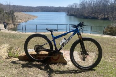 A blue Trek mountain bike is leaning against a rock in the foreground. In the background, a serene lake is visible, surrounded by bare trees and a pathway with a railing. A person can be seen walking along the path near the water. The scene is set on a sunny day with clear blue skies. Wyandotte County Lake - WyCo mountain bike trail.