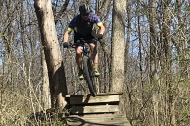 A mountain biker jumping off a wooden ramp in a forested area, surrounded by trees and greenery under a clear blue sky. The biker is wearing a helmet and colorful riding gear, focused on maintaining balance mid-air. Wyandotte County Lake - WyCo mountain bike trail.