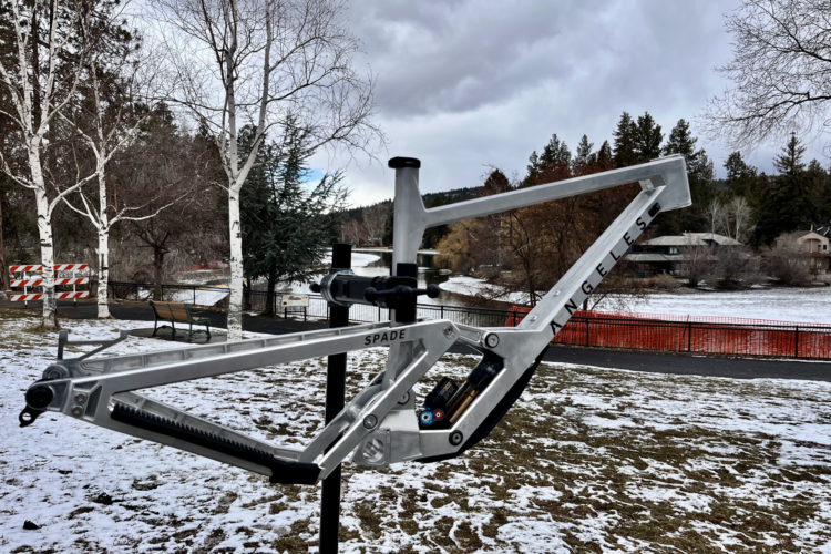 A silver bicycle frame labeled "SPADE" is mounted on a stand in a snowy park setting, with trees in the background and a river visible. The sky is overcast, creating a moody atmosphere.