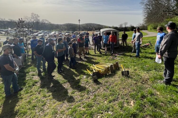 A group of people gathered in a grassy area, participating in an outdoor event. Some individuals are holding tools, while others listen to a speaker. The setting appears to be sunny, with trees in the background and a parking lot visible behind the group.