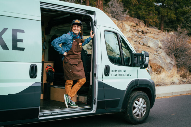 A smiling individual wearing a brown apron, denim shirt, and a cap stands in the doorway of a light green van with the word "BIKE" printed on the side. The van is parked in a natural setting with rocky terrain and sparse vegetation in the background. The person rests a hand on the van's door, exuding a welcoming and friendly demeanor.
