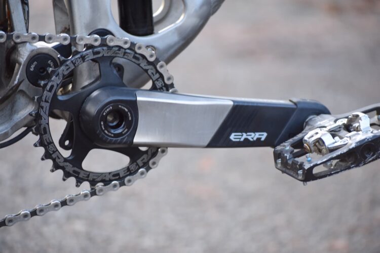 Close-up view of a bicycle crankset featuring a sleek black and silver design. The image showcases the pedal, crank arm, and chainring, along with a detailed look at the chain links. The crank arm is marked with the brand name "ERA," and the chainring has "RACEFACE" branding. The background is slightly blurred, emphasizing the components in focus.