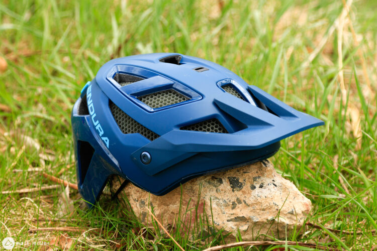 A blue mountain bike helmet resting on a rock, surrounded by green grass. The helmet features ventilation openings and a sleek design suitable for outdoor activities.