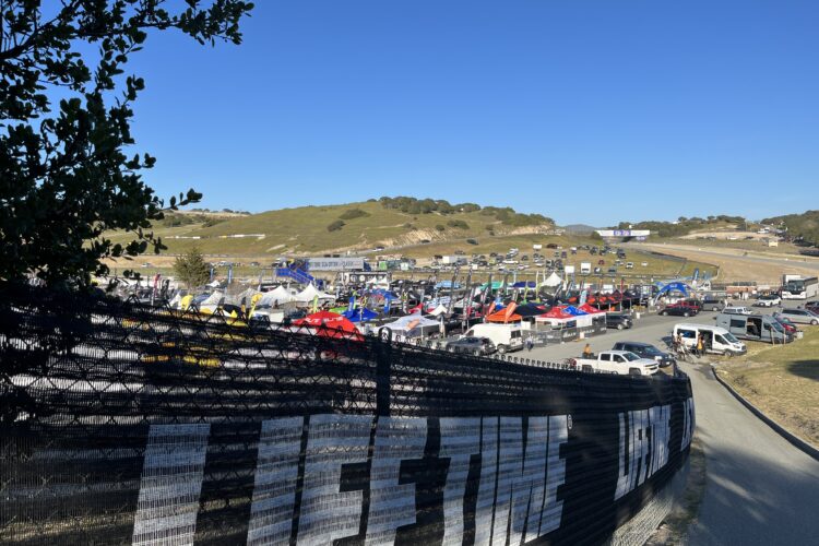 A panoramic view of a bustling outdoor event area with numerous colorful tents and vehicles, set against a clear blue sky. The scene features a large, black fence in the foreground displaying the word "LIFETIME," and rolling hills in the background with additional vehicles and event signage visible.