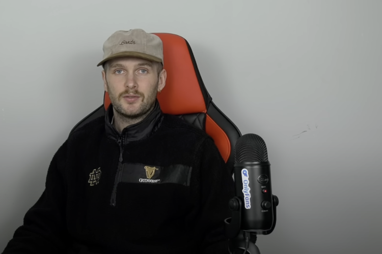 A person sitting in a gaming chair, wearing a beige cap and a black fleece sweatshirt with a Guinness logo. A microphone is positioned in front of them, and the background features a plain light-colored wall.