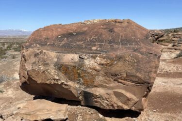 A large rock formation with ancient petroglyphs etched into its surface. The carvings are varied in design, showcasing geometric shapes and figures, set against a clear blue sky. In the background, a desert landscape stretches out, with distant mountains visible, partially covered in snow. Black Road Reboot mountain bike trail.
