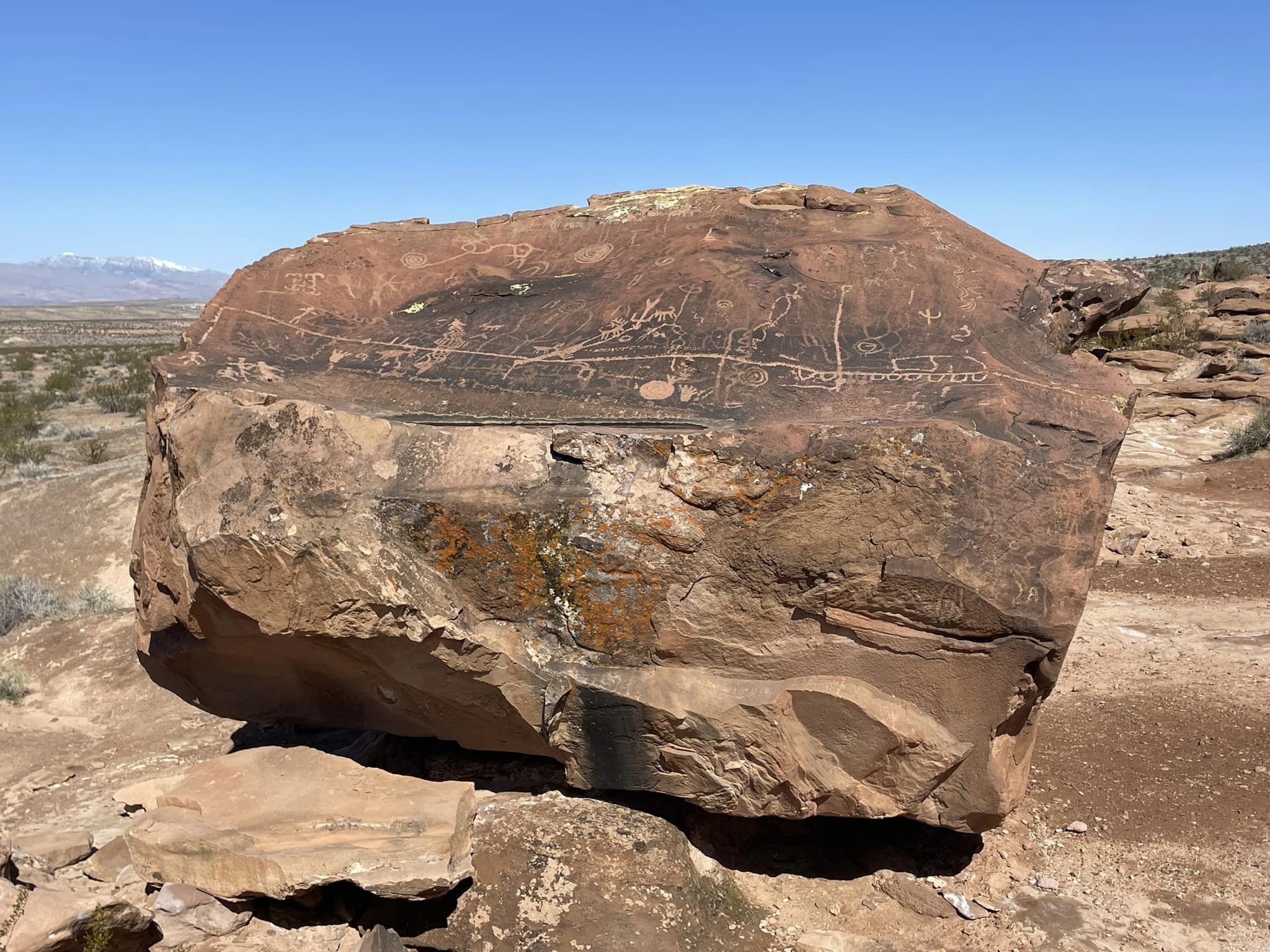 A large rock formation with ancient petroglyphs etched into its surface. The carvings are varied in design, showcasing geometric shapes and figures, set against a clear blue sky. In the background, a desert landscape stretches out, with distant mountains visible, partially covered in snow. Black Road Reboot mountain bike trail.