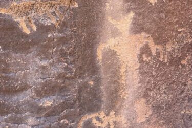 Ancient rock art depicting geometric shapes, animal figures, and human-like figures on a rugged stone surface. The petroglyphs showcase a range of designs, including a deer-like creature, and are rendered in earthy tones against the textured background of the rock. Black Road Reboot mountain bike trail.