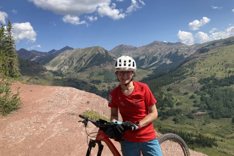 A person wearing a white helmet and glasses smiles while standing next to a mountain bike on a scenic mountain trail. They are dressed in a red shirt and blue shorts, with green forests and rugged mountains visible in the background under a partly cloudy sky.