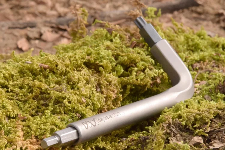 A silver hex wrench resting on green moss and leaves, with a blurred natural background.