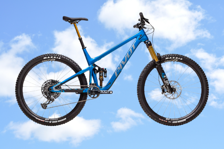A blue mountain bike suspended in front of a clear sky with fluffy white clouds. The bike features prominent components, including thick tires labeled "Maxxis," a sturdy frame, and a prominent suspension system.