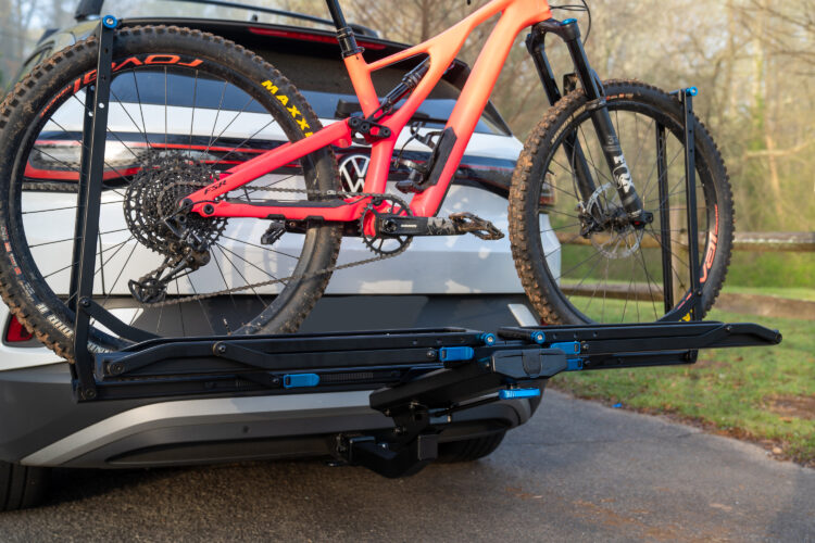 A close-up view of a mountain bike secured to a rear-mounted bike rack on a vehicle. The bike, featuring a bright orange frame and black tires with visible tread, is positioned upright with the rear wheel held in place by the rack. The scene includes part of the vehicle's rear, showcasing its modern design against a natural background.