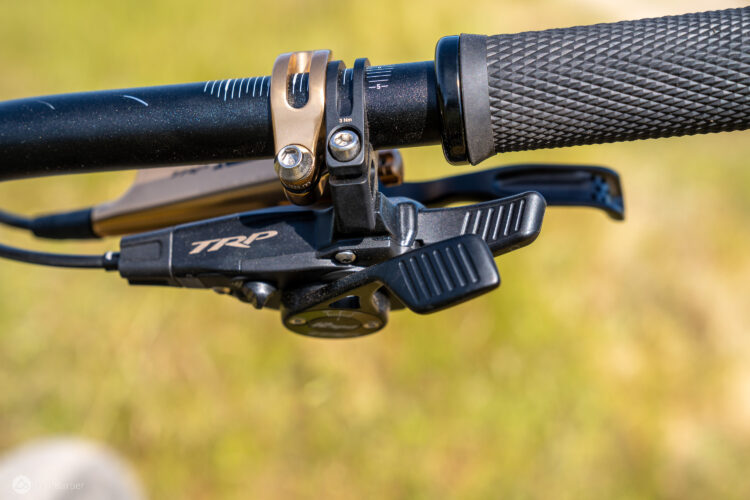 Close-up image of a black bicycle brake lever mounted on a handlebar, featuring a textured grip and a gold-colored clamp. The background is out of focus, highlighting the bike equipment.